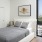 A bedroom with city view, featuring a white upholstered bed, gray quilt, and abstract art.