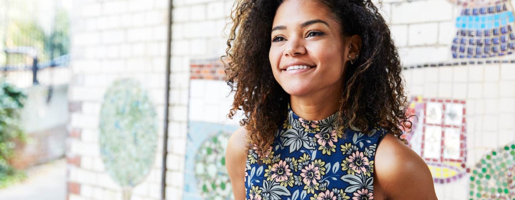 Smiling woman with curly hair wearing a floral top standing in front of a mosaic wall.