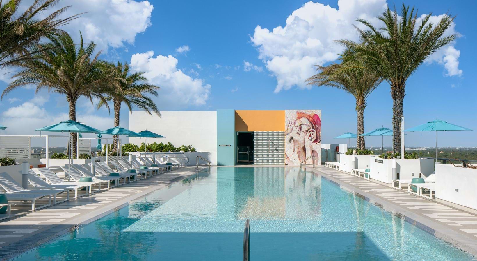 Sun-soaked relaxation meets elevated outdoor living Rooftop swimming pool with lounge chairs, blue umbrellas, palm trees, and colorful mural art.